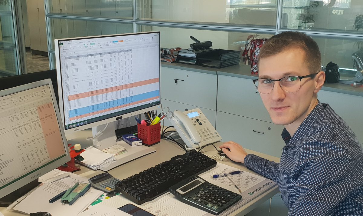 Loris at his workplace at Pepperl+Fuchs. There are two screens on his desk showing various tables. There are various office supplies such as documents, calculators, etc. on his desk.