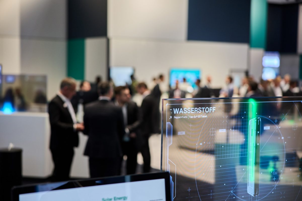 View of the Pepperl+Fuchs booth at the Hannover Messe 2023. In the blurred background, visitors to the Pepperl+Fuchs booth and Pepperl+Fuchs employees can be seen standing and talking together. In the sharp foreground, an exhibit is visible.