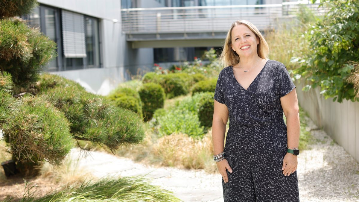 Kristen, Marketing Manager Americas at Pepperl+Fuchs Twinsburg, stands in an area planted with shrubs in front of the Pepperl+Fuchs building in Mannheim and smiles into the camera.