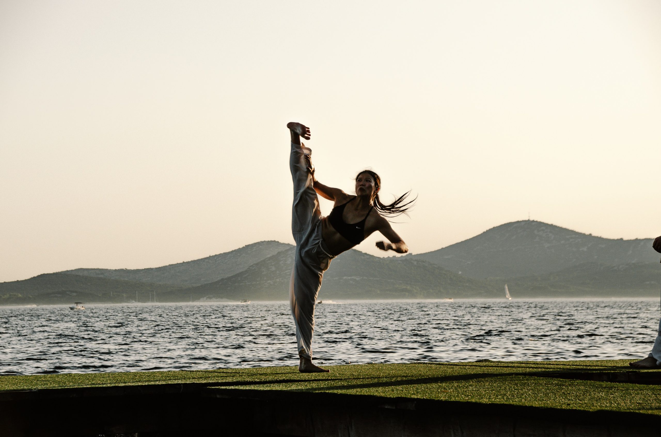 Yun Xia is standing in a meadow. Behind her is a lake and a hilly landscape. She stretches one leg up in the air, as if doing the splits.