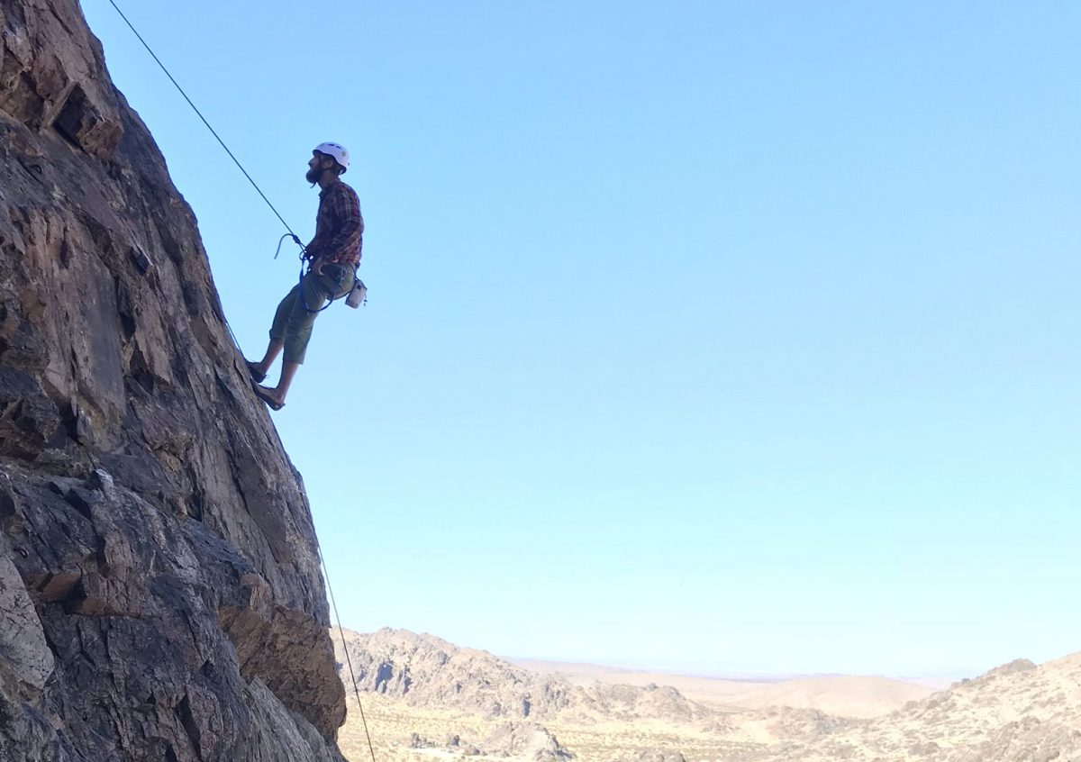 Lucas climbing in the mountains. It hangs securely on a wall. It is blue sky, the sun is shining and in the background you can see far over the mountains.