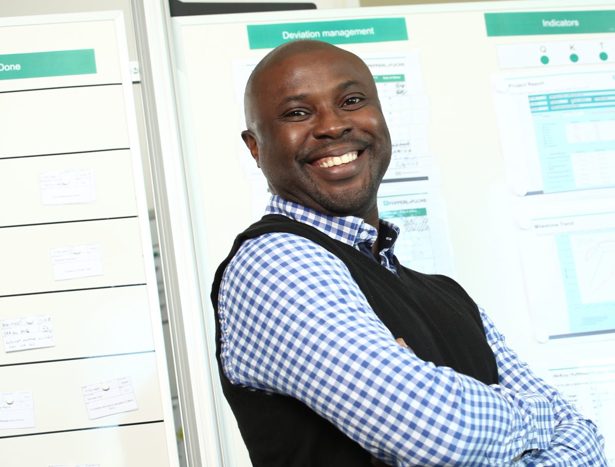 Obi stands in front of various flipcharts with various plans hanging on them. He has his arms crossed and is smiling at the camera.
