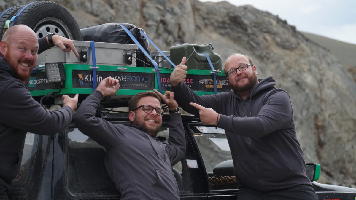 The three colleagues in front of their car. They give their thumbs up or point to their packed car.