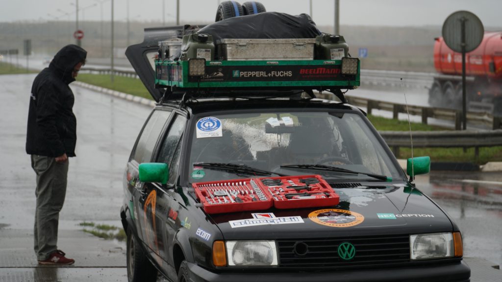 View of the car from the front. A toolbox lies on the hood. A man stands at the back of the car and looks towards the open trunk. It's raining.