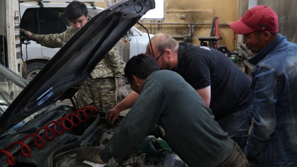 In the workshop, three men lean over the open hood.