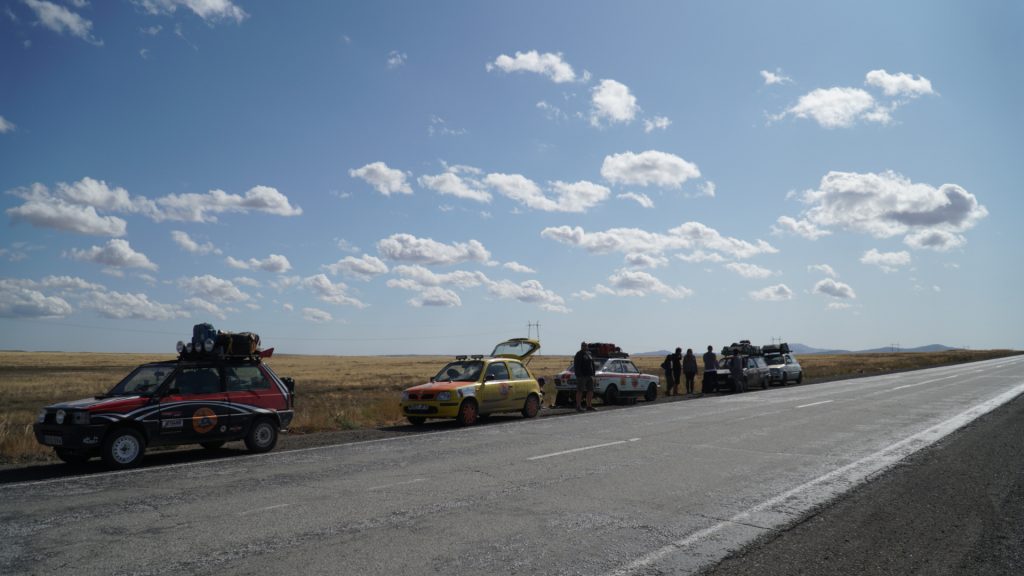 There are four cars on the side of the road. The drivers talk. It is blue sky and the sun is shining.