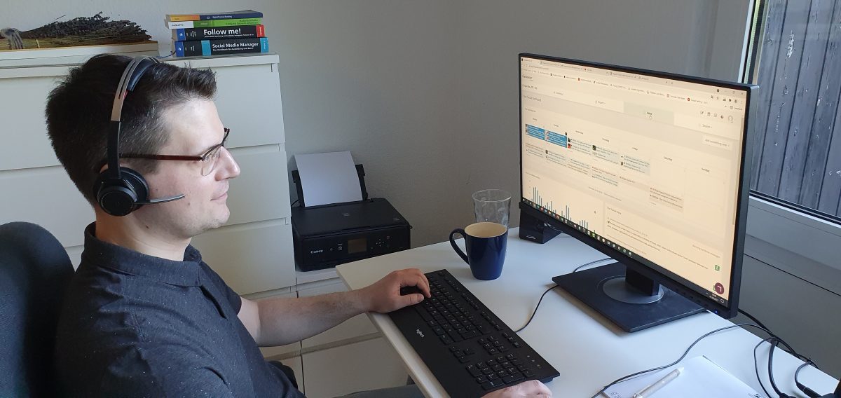 Matthias, Social Media Manager at Pepperl+Fuchs, at his home office. He is wearing a headset and looking at his screen. Behind him is a white chest of drawers with specialist literature on it.