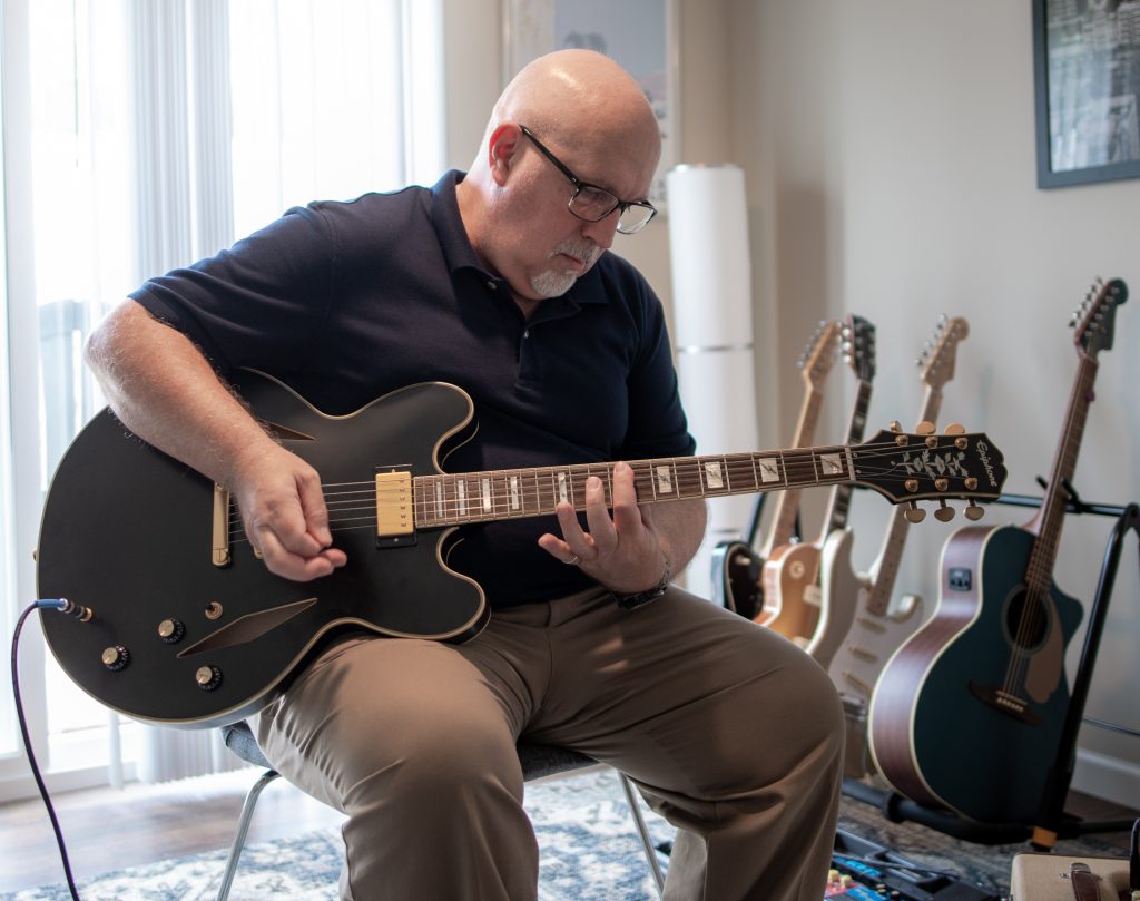 Andrew, Sales Performance Coach at Pepperl+Fuchs, sits on a chair in a room and plays the guitar. In the background you can see several guitars standing on a guitar stand.