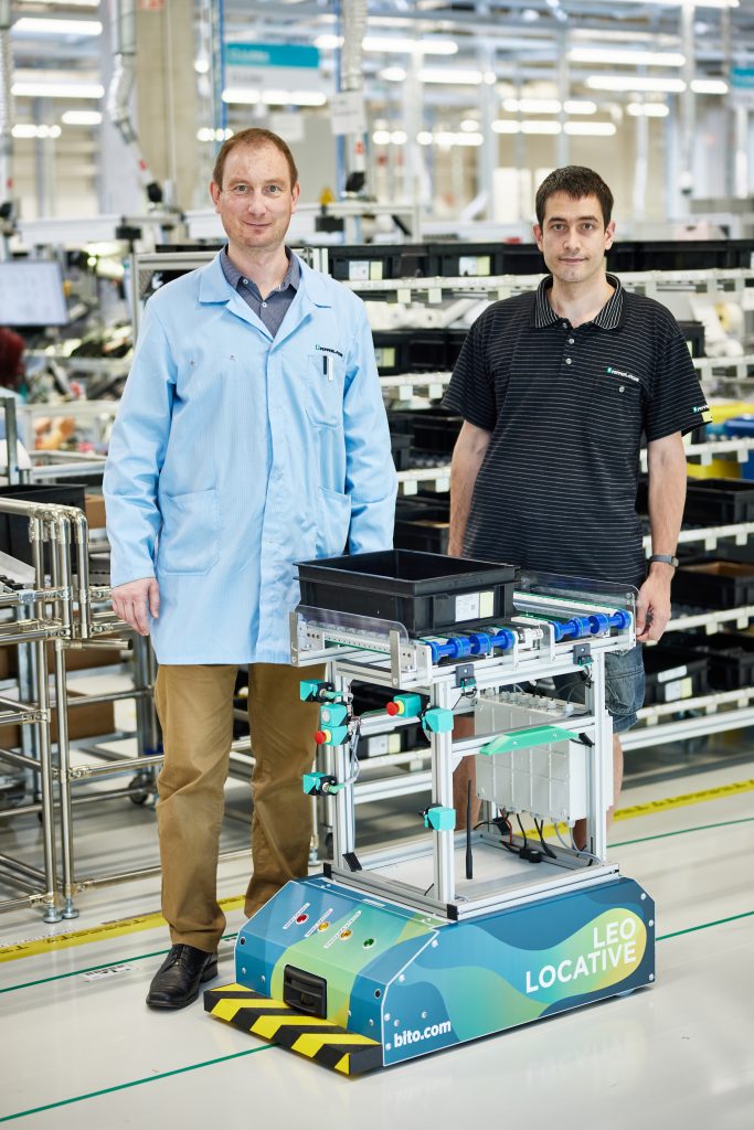 Lean consultant Stepan and Pavel, test support in the electromagnetic sensors (EMS) product area Pavel at Pepperl+Fuchs in the Czech Republic, stand in the production hall and smile into the camera. In front of them is the LEO-Locative transport system.