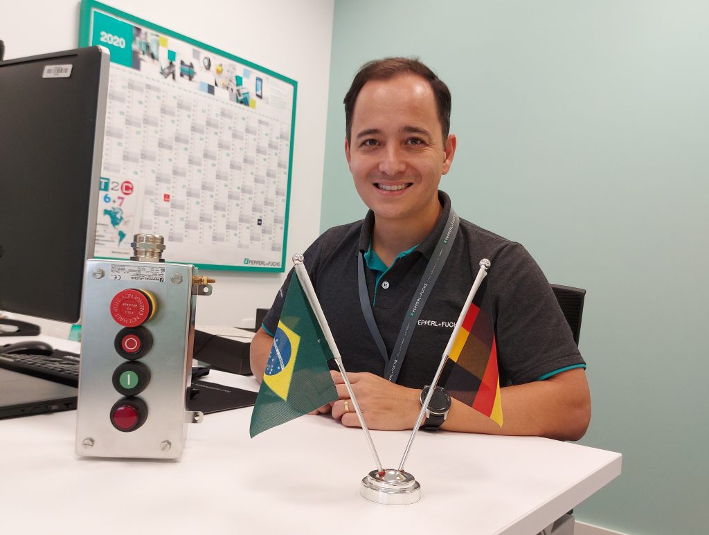 Fabio, Regional Sales Director PA at Pepperl+Fuchs in Brazil, sits at his desk and smiles into the camera. His screen is slightly offset to the left in front of him. Directly in front of him are a German and a Brazilian table flag.