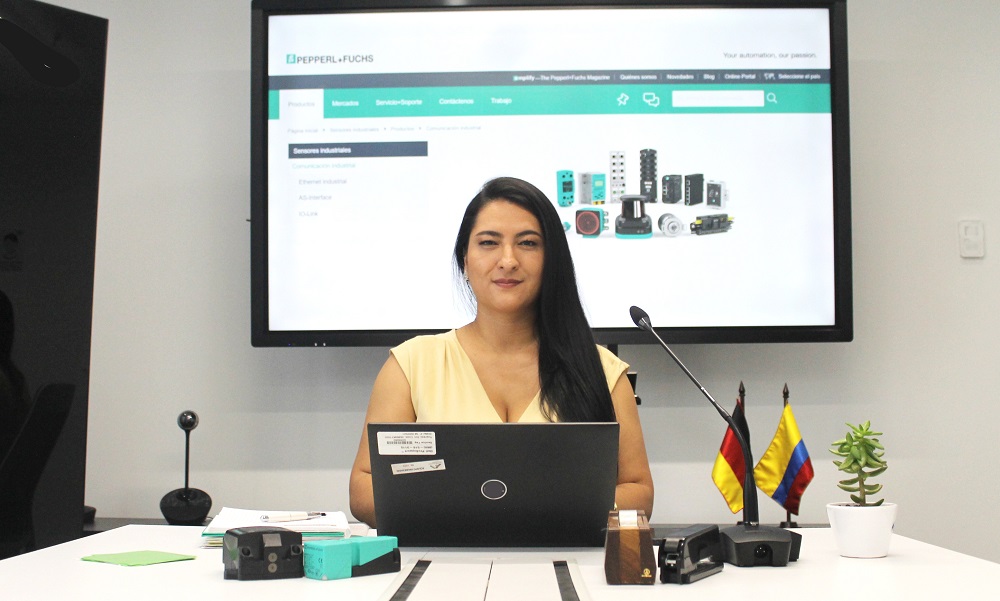 Marcela sits at her desk at her workplace at Pepperl+Fuchs and looks into the camera. Her laptop is open in front of her. Behind her you can see a video screen on the wall showing the Pepperl+Fuchs company website. There are also office supplies on the table, such as a stapler and a hole punch. Furthermore, to the right of her there is a small plant as well as a German and a Colombian table flag.