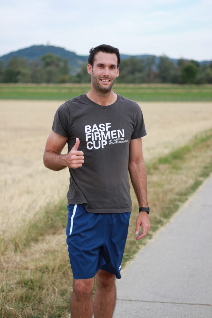 André, Product Compliance Engineer at Pepperl+Fuchs, is standing on a dirt path wearing a running shirt and running shorts. He looks into the camera and raises his right thumb up.