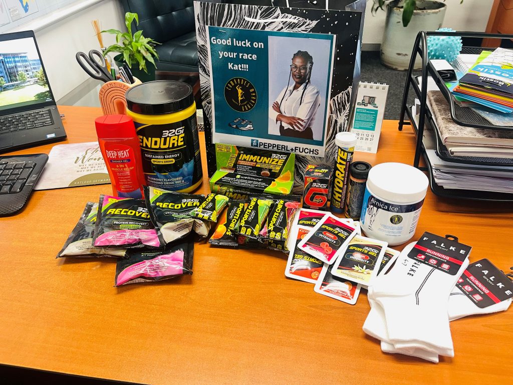 Katlegos' desk at Pepperl+Fuchs. On her desk, there are various dietary supplements, such as bars, powder packets, a powder container, as well as a jar of Anika's ointment and socks. In the center, there is card with a photo of Katlego and the phrase "Good luck on your race Kat!!!"