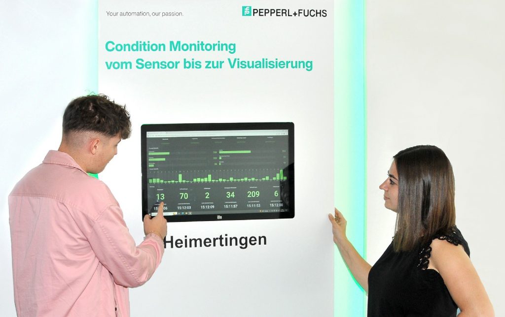 In the middle of the image, you can see the dashboard. The display shows the levels of the smart coffee machine. To the left of the display, a man is gesturing with his right hand, explaining one of the values. To the right of the display, a woman is looking interestedly at the display and listening to the man.