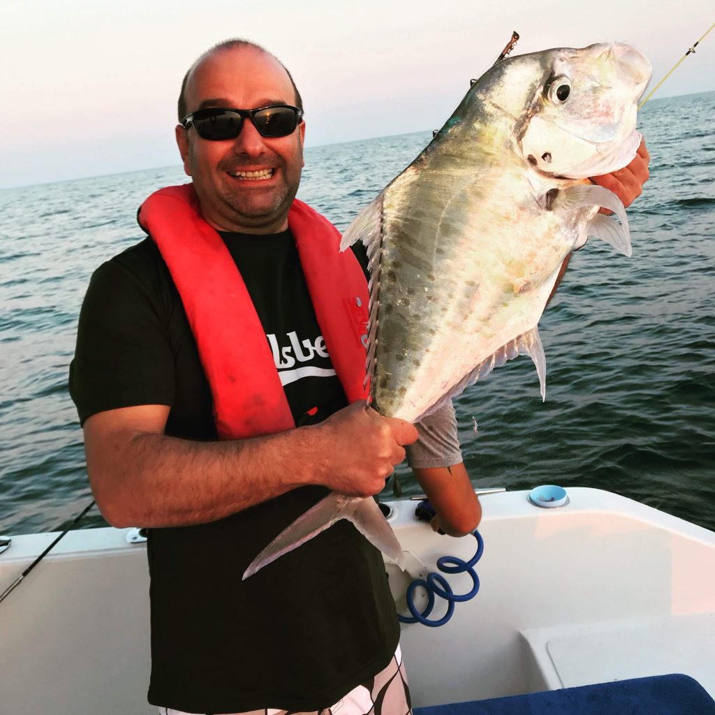 Stefano, Sales Director at Pepperl+Fuchs in Sulbiate, stands on a boat wearing a shirt, shorts, and sunglasses, smiling as he presents his large fish that he has caught with both hands in front of him to the camera.