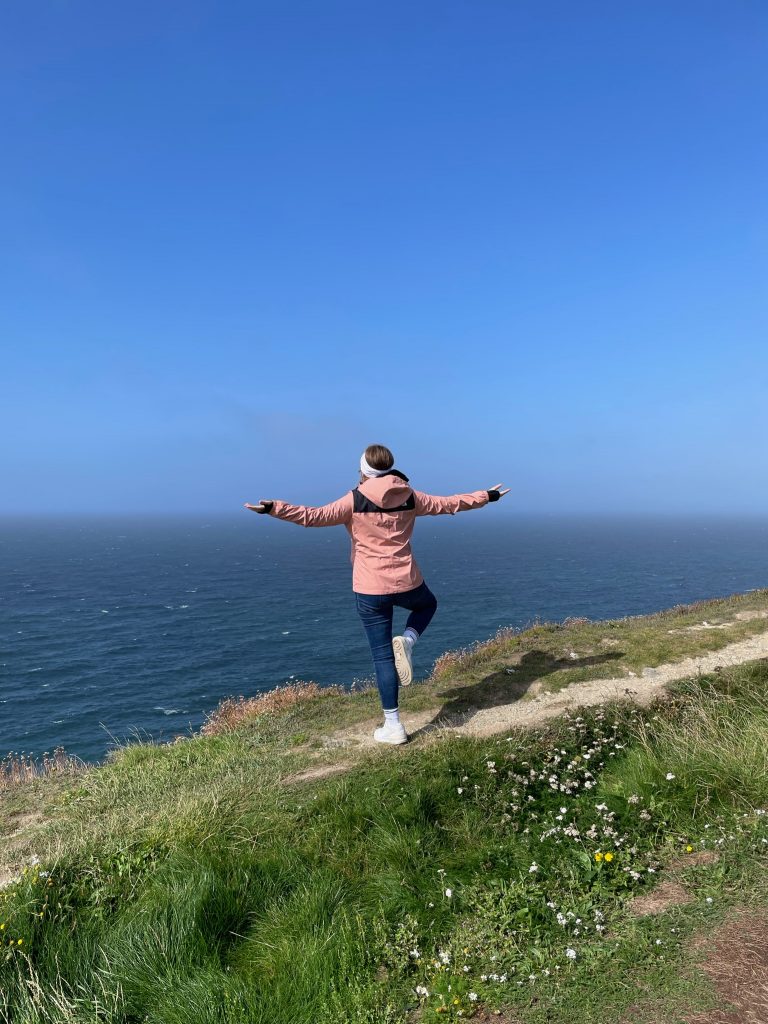 Anna-Lena is standing on a small path with her back to the camera. She is looking at the sea, her arms stretched out and one leg bent.