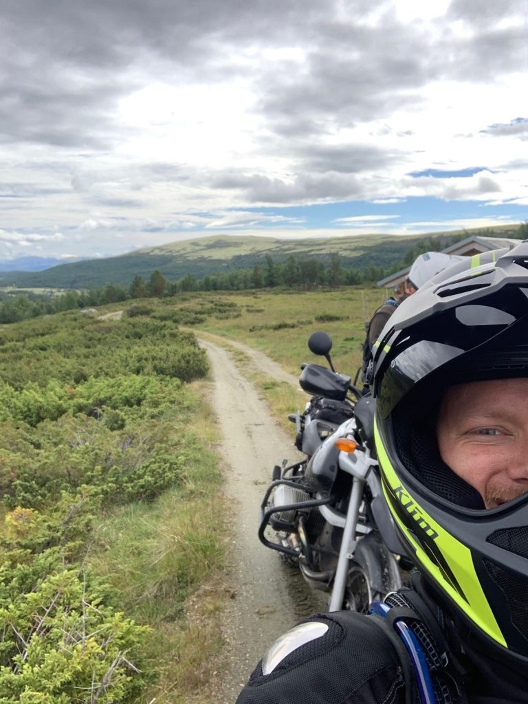 Robert is standing on a narrow, dirt road winding through a green, hilly landscape. It's cloudy. He's standing in front of his motorcycle, taking a selfie. He's wearing motorcycle gear and smiling at the camera.