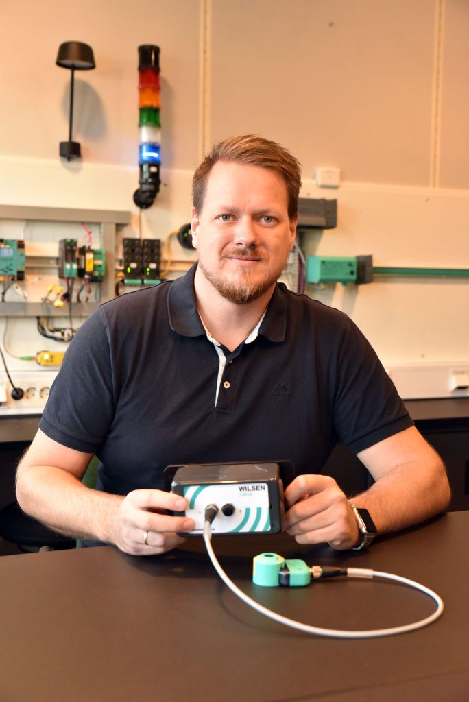 Robert, System & Solution Engineer, at his workstation. He sits at the table with the Wilsen sensor in front of him. Various devices can be seen behind him.