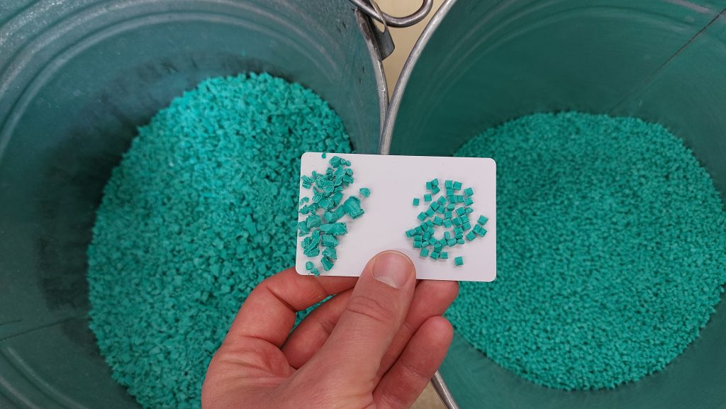 Two bins are visible. The left bin contains the ground material (regrind), and the right bin contains the new granulate. A hand can be seen holding a small, white box on which the two materials can be seen closer together. The material is turquoise in color.