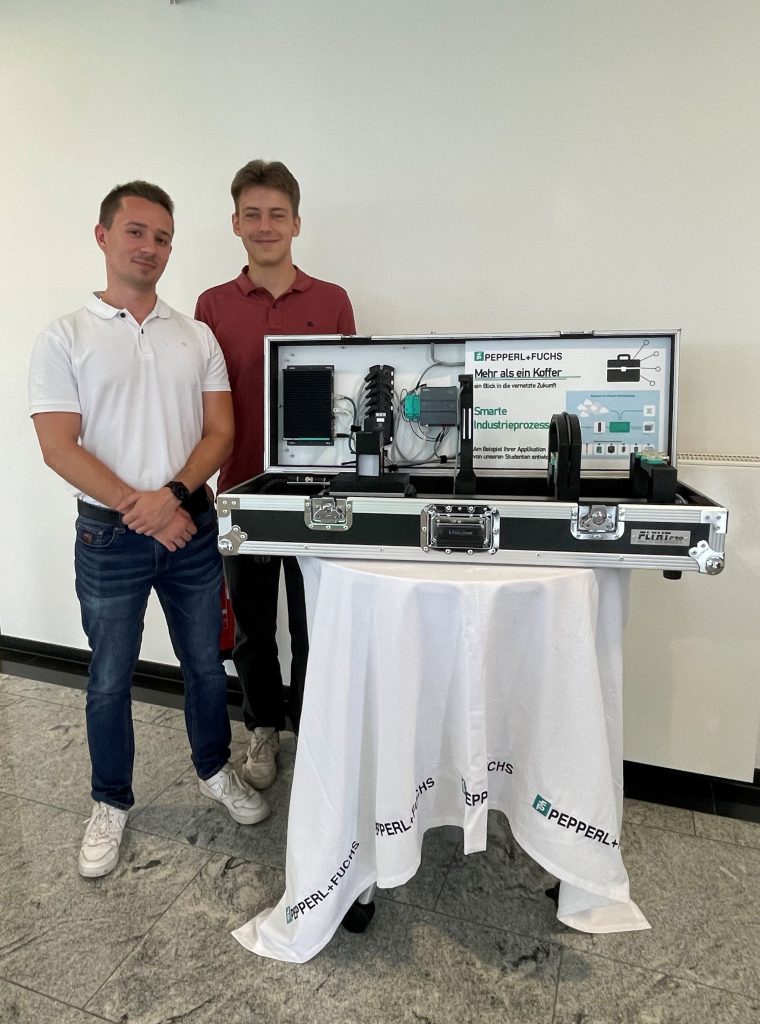 Practical phases at Pepperl+Fuchs: Daniel and David stand behind the open case exhibit, which sits on a round table covered with a white tablecloth bearing the Pepperl+Fuchs logo. The built-in sensors can be seen inside.