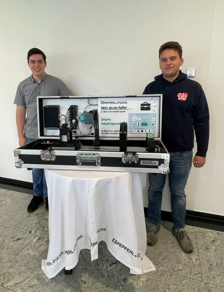 Practical phases at Pepperl+Fuchs: Dennis and Aaron stand behind the open case exhibit, which sits on a round table covered with a white tablecloth bearing the Pepperl+Fuchs logo. The built-in sensors can be seen inside.