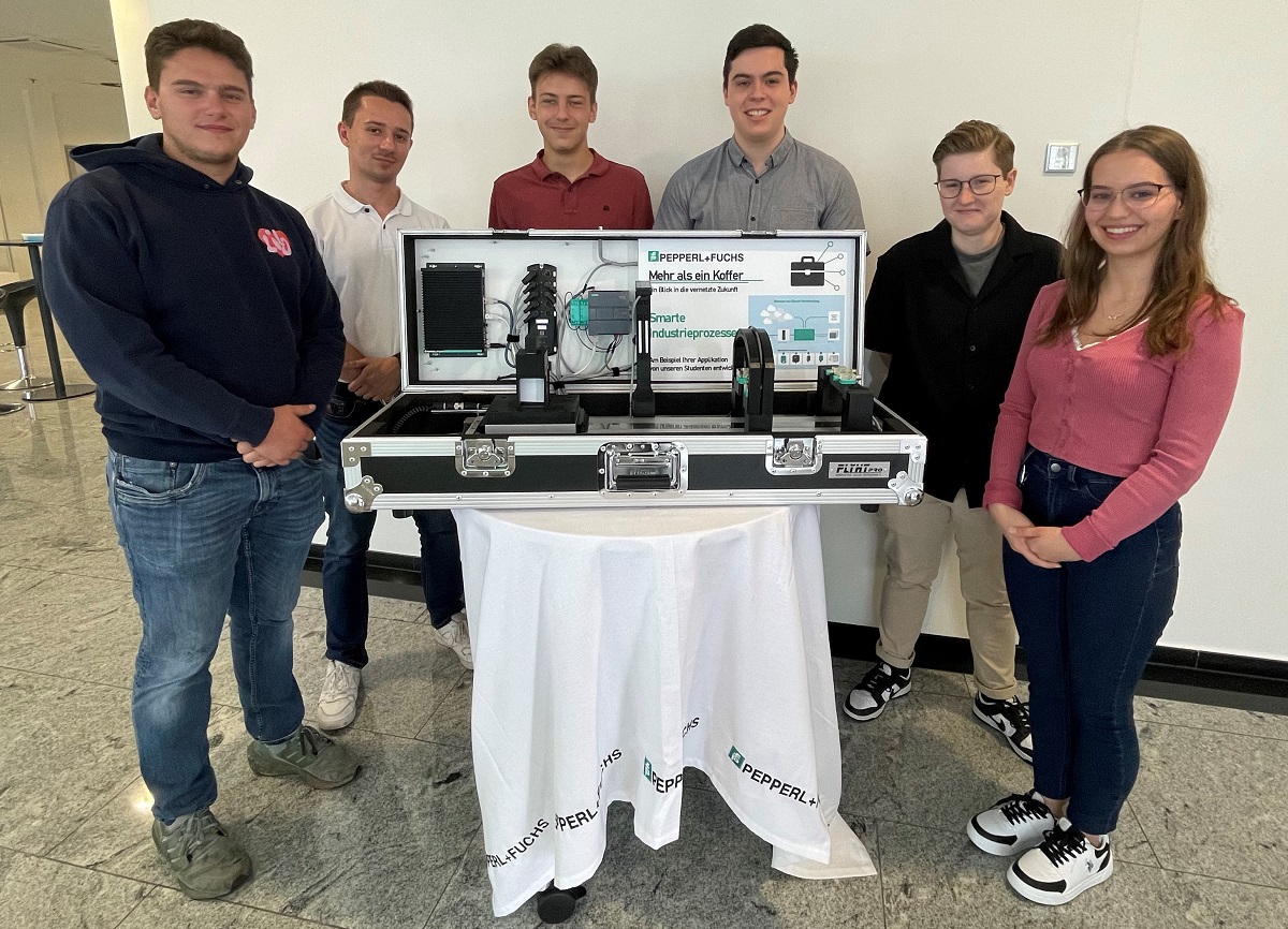 Six dual study students stand around a round table covered with a white tablecloth, on which the finished suitcase exhibit rests. They all look into the camera and smile.
