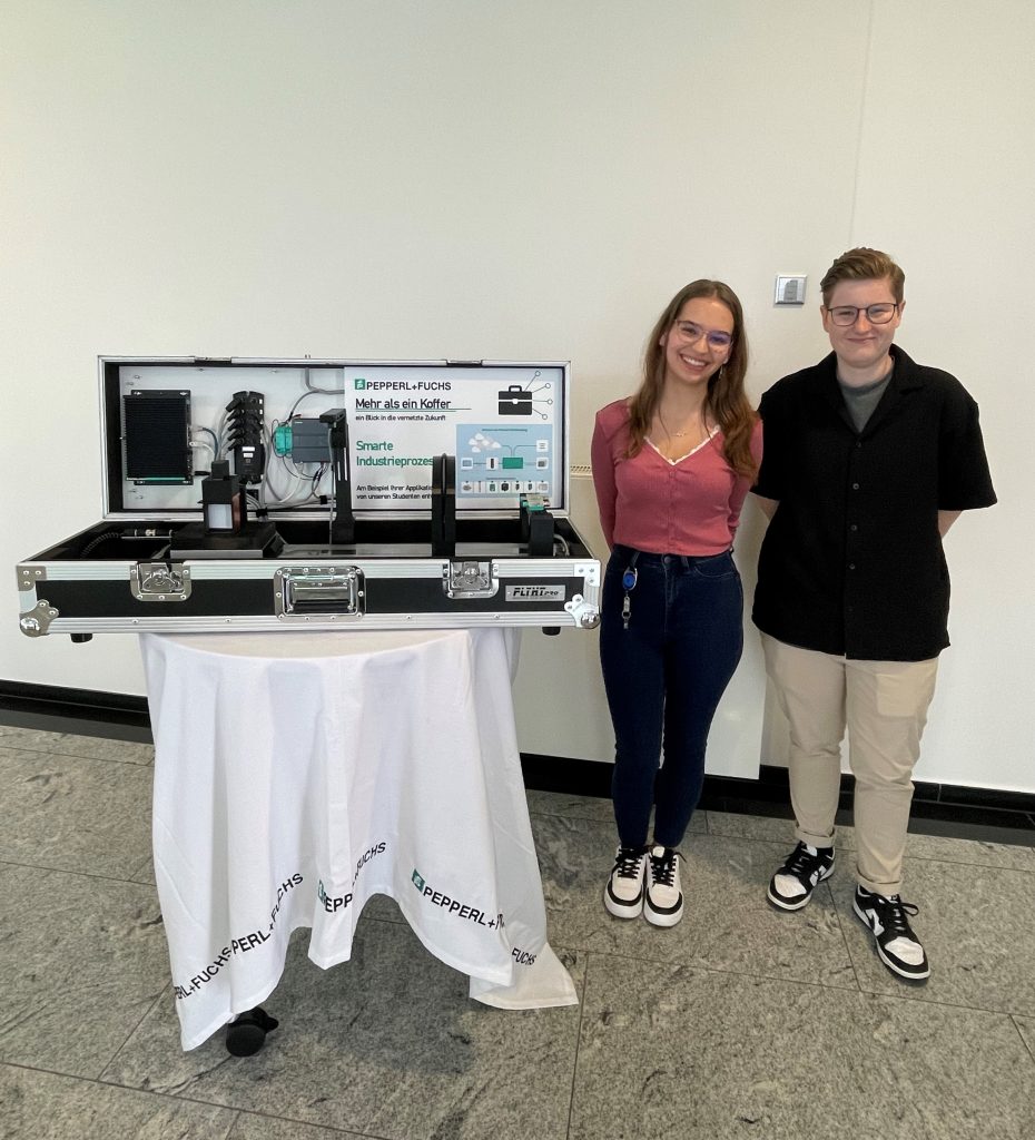Practical phases at Pepperl+Fuchs: Laura and Alina stand behind the open case exhibit, which sits on a round table covered with a white tablecloth bearing the Pepperl+Fuchs logo. The built-in sensors can be seen inside.