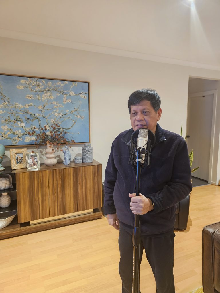 Benjamin, Quality & Compliance Manager, is standing in his living room at home, singing into a microphone. Behind him is a sideboard with personal pictures and a large painting on the wall.
