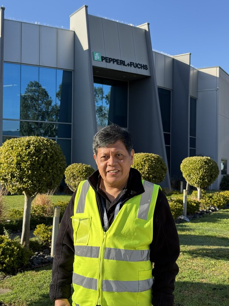 Benjamin, Quality & Compliance Manager, stands in front of the Pepperl+Fuchs headquarters in Melbourne. He's wearing a yellow safety vest and smiling at the camera.