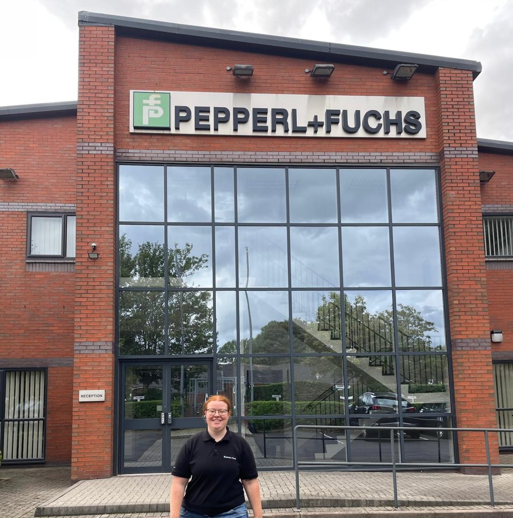 Melanie completed a semester abroad in Oldham. Here she is standing in front of the entrance to the Pepperl+Fuchs building in Oldham. It's a red brick building with a large glass frontage directly behind her.