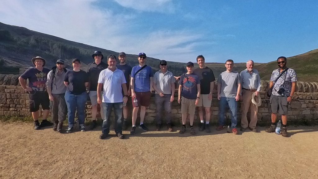 Department photo at Dovestone Reservoir: The 13 colleagues stand side by side in front of a small wall, with a hilly landscape and blue sky in the background.