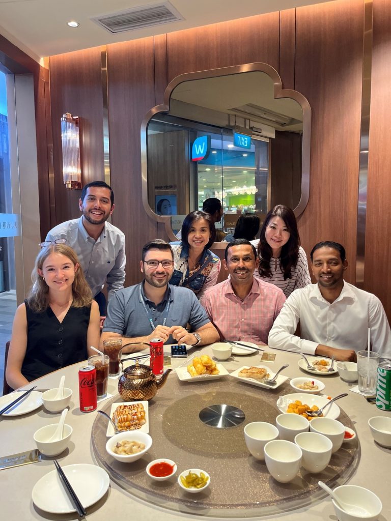Janine with her six colleagues. Four of them are sitting at a round table, and three are standing behind them. Everyone is smiling at the camera.