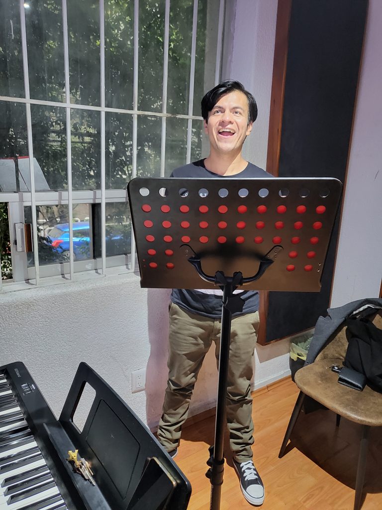 Daniel is standing in a small room in front of a music stand and is singing. A keyboard can be seen to his left.